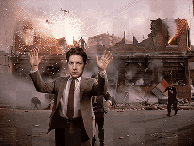 A Man In A Suit And Tie Is Standing In Front Of A Destroyed Building GIF
