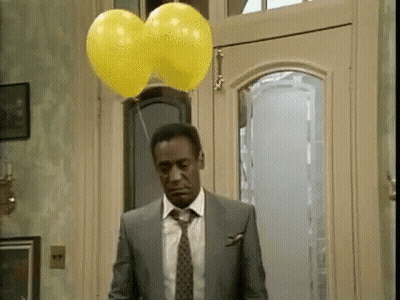 A Man In A Suit And Tie Is Standing In Front Of A Door With Two Yellow Balloons Hanging From It . GIF