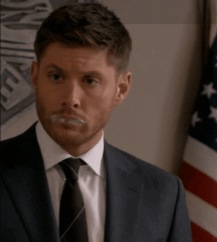A Man In A Suit And Tie Is Standing In Front Of A Flag GIF