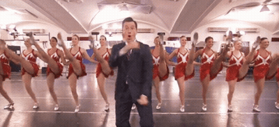 A Man In A Suit And Tie Is Standing In Front Of A Group Of Cheerleaders GIF