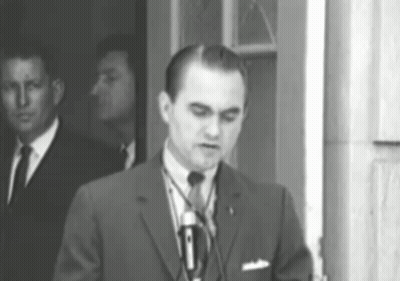 A Man In A Suit And Tie Is Standing In Front Of A Microphone GIF