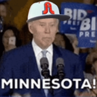 A Man In A Suit And Tie Is Standing In Front Of A Microphone Wearing A Baseball Cap That Says Minnesota . GIF