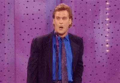 A Man In A Suit And Tie Is Standing In Front Of A Purple Curtain And Pointing At Something . GIF