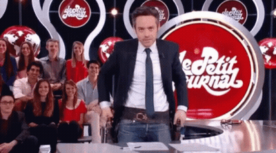 A Man In A Suit And Tie Is Standing In Front Of A Red Sign That Says Le Petit Journal GIF