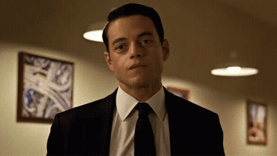 A Man In A Suit And Tie Is Standing In Front Of A Wall With Pictures On It GIF