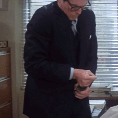 A Man In A Suit And Tie Is Standing In Front Of A Window With Blinds That Says ' A ' On It GIF