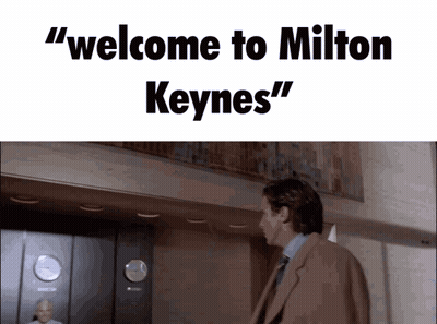A Man In A Suit And Tie Is Standing In Front Of An Elevator With The Words " Welcome To Milton Keynes " Above Him GIF