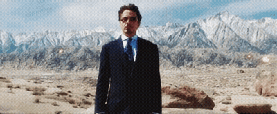 A Man In A Suit And Tie Is Standing In The Desert With Mountains In The Background GIF