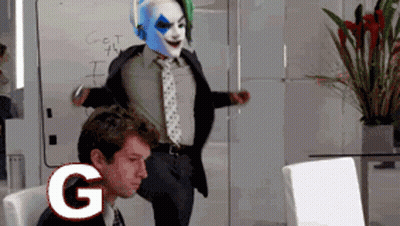 A Man In A Suit And Tie Is Standing Next To A Clown In A Mask With The Letter G Above Him GIF