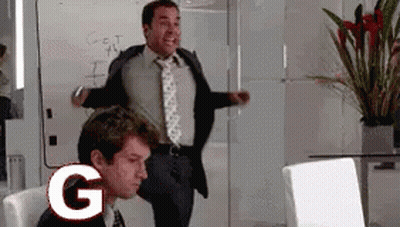 A Man In A Suit And Tie Is Standing Next To A Man In A Chair With The Letter G In Front Of Him GIF