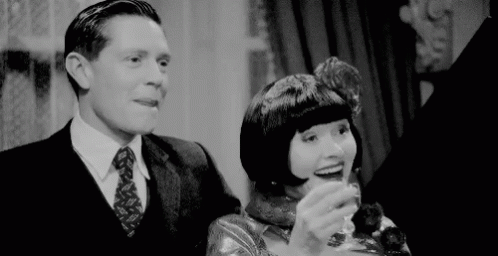 A Man In A Suit And Tie Is Standing Next To A Woman Holding A Glass Of Wine . GIF