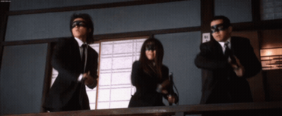 A Man In A Suit And Tie Is Standing Next To A Woman In A Mask And Holding A Gun GIF