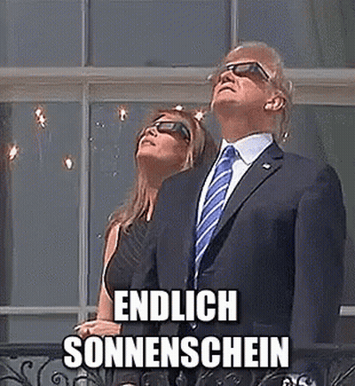 A Man In A Suit And Tie Is Standing Next To A Woman Wearing Sunglasses And Looking Up At The Sun . GIF