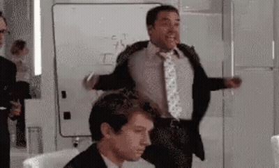 A Man In A Suit And Tie Is Standing Next To Another Man In A Chair . GIF
