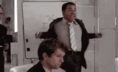A Man In A Suit And Tie Is Standing Next To Another Man In A Chair . GIF