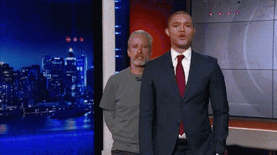 A Man In A Suit And Tie Is Standing Next To Another Man In A Grey Shirt GIF