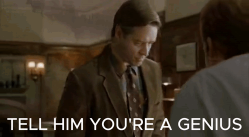 A Man In A Suit And Tie Is Standing Next To Another Man In A Living Room And Talking To Him . GIF