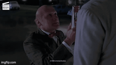 A Man In A Suit And Tie Is Standing Next To Another Man With The Words Slacker On The Screen GIF