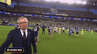 A Man In A Suit And Tie Is Standing On A Soccer Field With A Referee View Button On The Bottom GIF