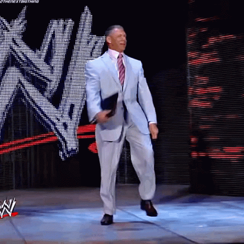 A Man In A Suit And Tie Is Standing On A Stage In Front Of A Wwe Logo GIF