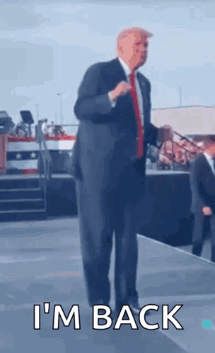 A Man In A Suit And Tie Is Standing On A Stage With The Words I 'M Back Written Below Him . GIF