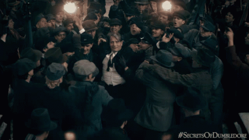 A Man In A Suit And Tie Is Surrounded By A Crowd With The Hashtag Secrets Of Dumbledore On The Bottom GIF
