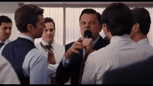 A Man In A Suit And Tie Is Talking Into A Microphone While Another Man Looks On GIF