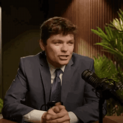 A Man In A Suit And Tie Is Talking Into A Microphone While Holding A Coffee Mug With The Letter Af On It . GIF