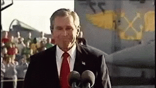 A Man In A Suit And Tie Is Talking Into Two Microphones . GIF
