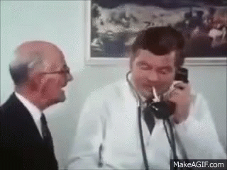 A Man In A Suit And Tie Is Talking On A Phone While Another Man Laughs . GIF