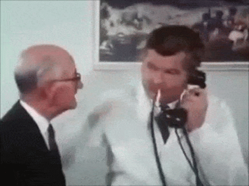A Man In A Suit And Tie Is Talking On A Phone While Another Man Looks On . GIF