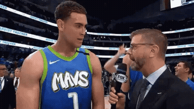 A Man In A Suit And Tie Is Talking To A Basketball Player In A Blue And Green Jersey . GIF