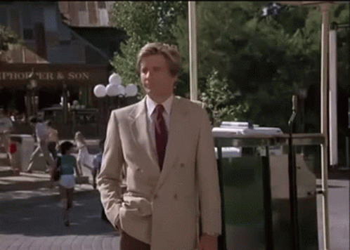 A Man In A Suit And Tie Is Walking Down A Street In Front Of A Store Called Thompson And Son GIF