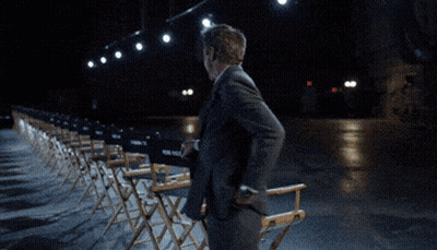 A Man In A Suit And Tie Is Walking In Front Of A Row Of Chairs GIF