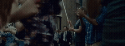 A Man In A Suit And Tie Is Walking Through A Crowd Of People Holding A Stick . GIF