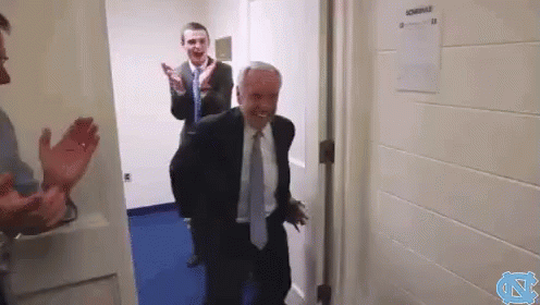 A Man In A Suit And Tie Is Walking Through A Doorway While A Man Applauds Behind Him . GIF