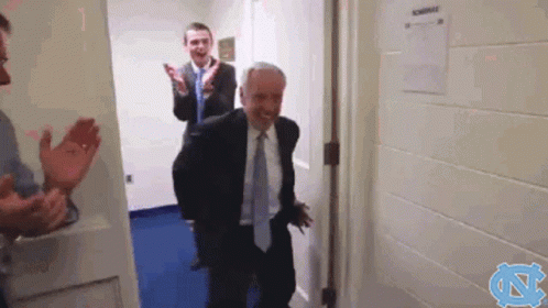A Man In A Suit And Tie Is Walking Through A Doorway While Another Man Applauds Behind Him GIF