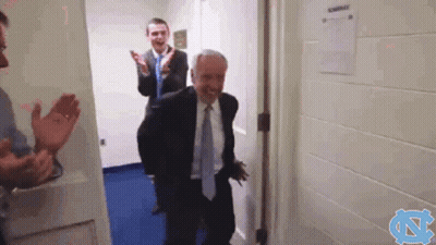 A Man In A Suit And Tie Is Walking Through A Doorway While Another Man Applauds . GIF