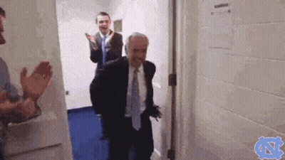 A Man In A Suit And Tie Is Walking Through A Doorway While Other Men Applaud . GIF