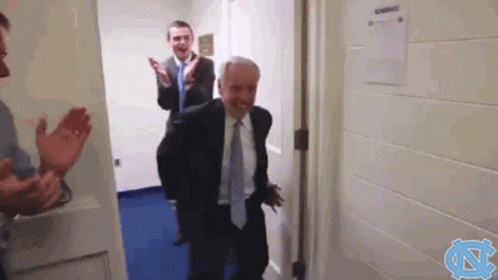 A Man In A Suit And Tie Is Walking Through A Doorway While Two Other Men Applaud Behind Him . GIF