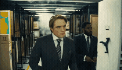 A Man In A Suit And Tie Is Walking Through A Hallway With A Box That Says Fragile On It GIF