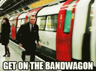 A Man In A Suit And Tie Is Walking Towards A Train With The Words Get On The Bandwagon Below Him GIF