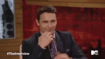 A Man In A Suit And Tie Is Waving His Hand While Sitting On A Stage . GIF
