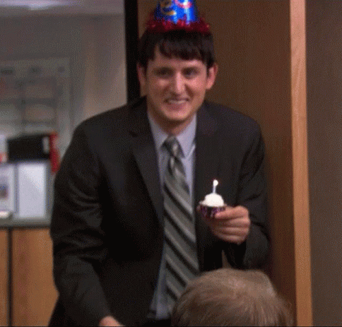 A Man In A Suit And Tie Is Wearing A Party Hat And Holding A Cupcake With A Candle In It . GIF