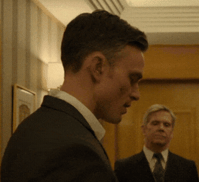 A Man In A Suit And Tie Looks Down While Another Man Stands Behind Him GIF