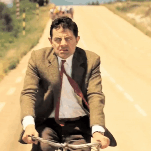 A Man In A Suit And Tie Rides A Bike Down A Dirt Road GIF