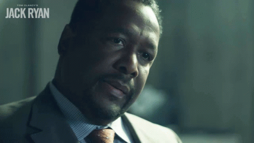 A Man In A Suit And Tie Says Okay In Front Of A Poster For Tom Clancy 'S Jack Ryan GIF