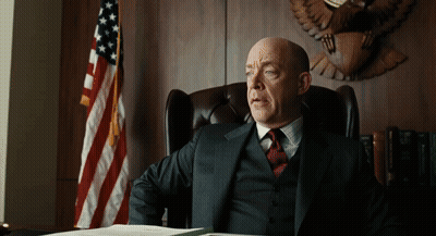 A Man In A Suit And Tie Sits At A Desk In Front Of An American Flag GIF
