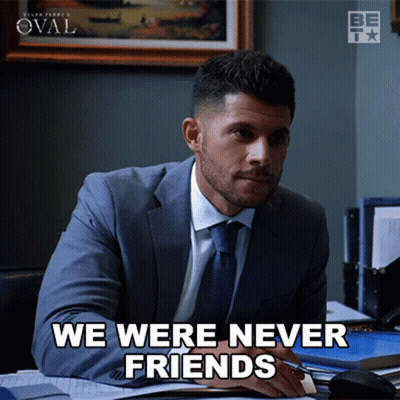 A Man In A Suit And Tie Sits At A Desk With The Words We Were Never Friends Below Him GIF