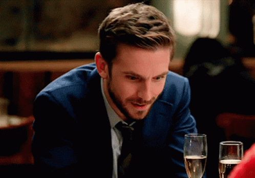 A Man In A Suit And Tie Sits At A Table With A Glass Of Champagne GIF